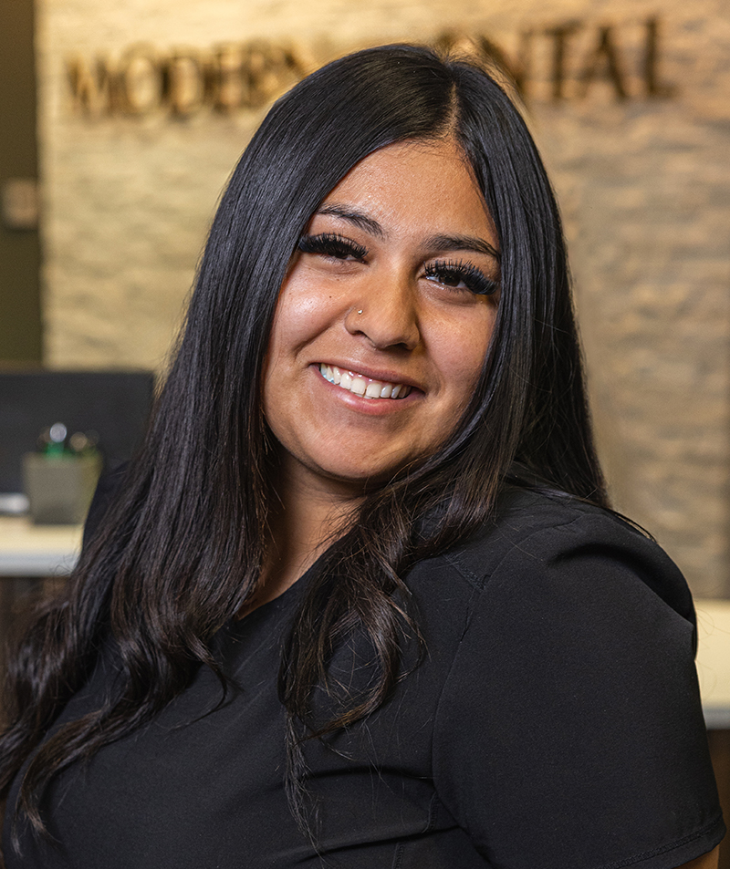 Professional woman smiling at dental office, wearing black shirt against blurred clinic background.
