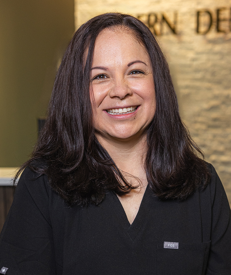Professional woman with long dark hair smiling in dental office setting wearing black scrubs.