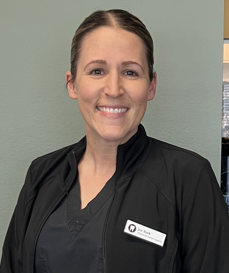 Professional dental staff member in black scrubs smiling at camera in clinical setting.