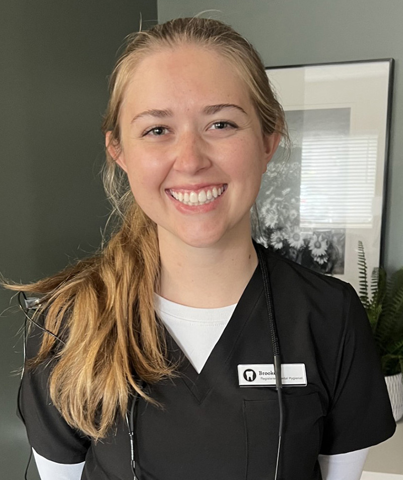 Young dental professional smiling at camera wearing black scrubs in modern dental office setting.