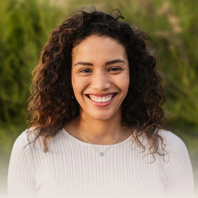 Smiling woman with curly dark hair wearing light sweater outdoors with green background.